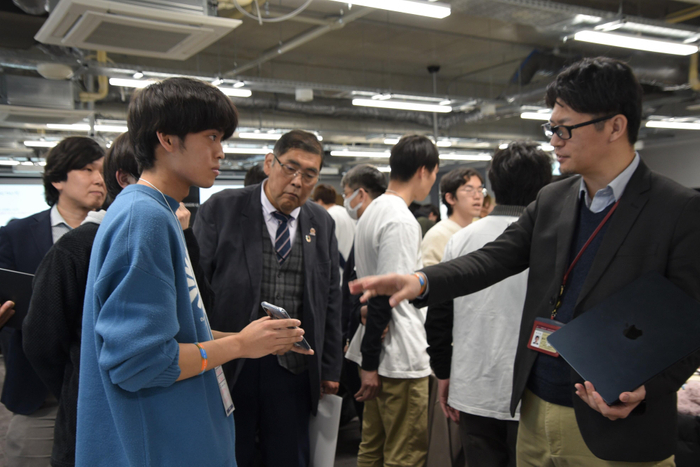 学生の説明を聞く小原学長や情報工学部の鈴木秀和准教授（右）