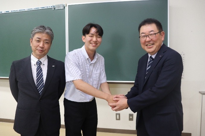 がっちり握手して藤原さん(中央)を激励する烏田社長(右)。左は小林マネージャー