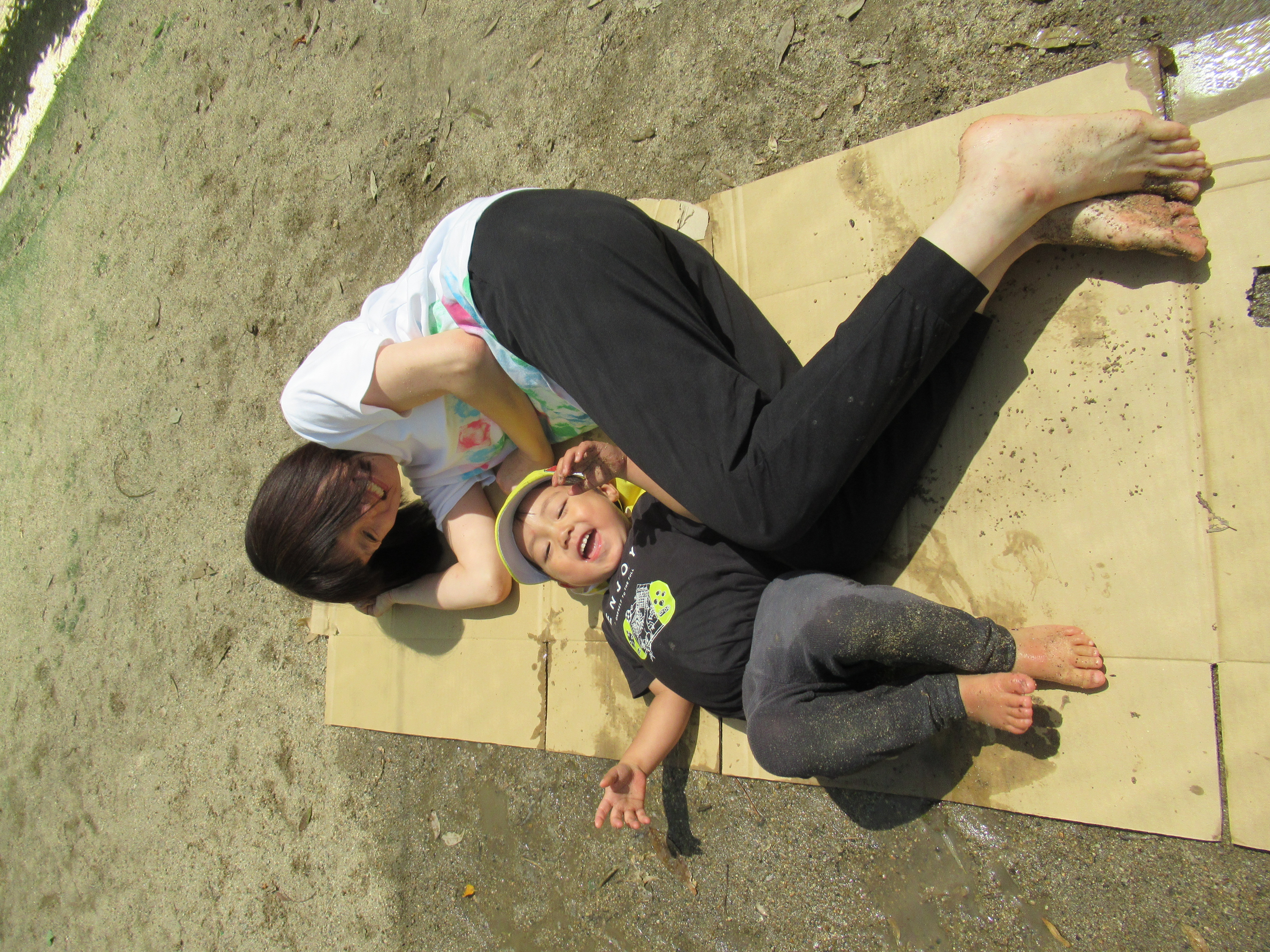 泥のついたダンボールの上に寝転ぶ園児と保育者